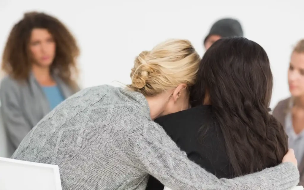 Looking at the back of two women sitting in a group. One woman is leaning on another woman and sad.